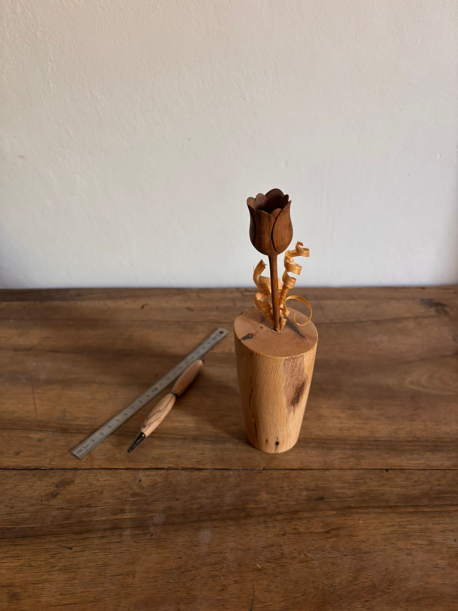 Une fleur de tulipe sculptée en bois brun posée sur un socle en bois brut, avec deux fins copeaux de bois enroulés.