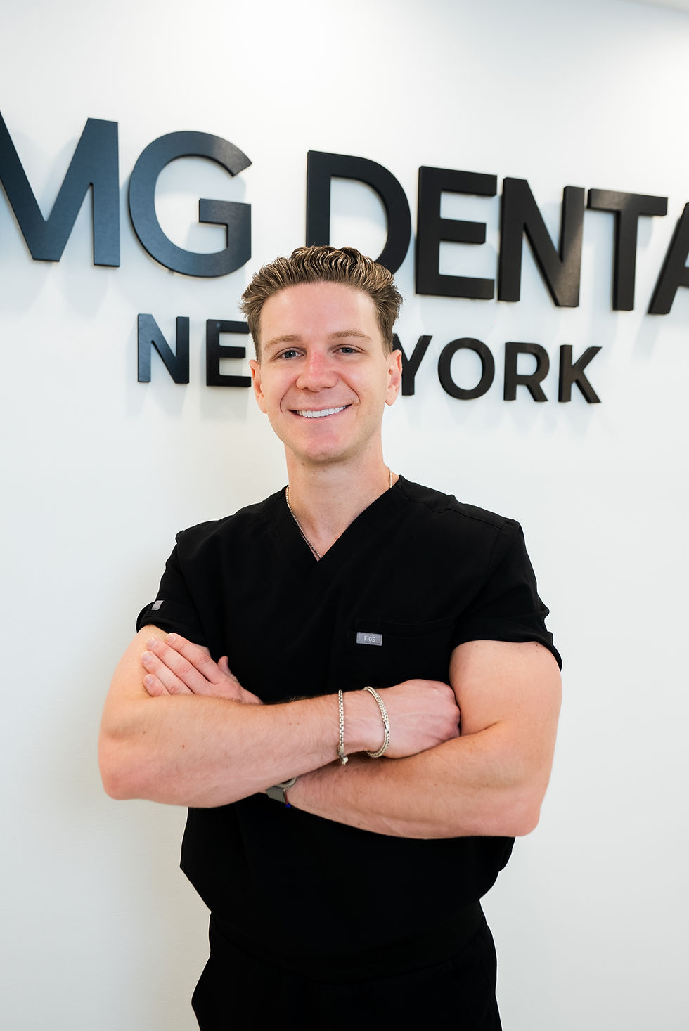 Dentist in black scrubs standing with arms crossed in front of the AMG Dental New York logo.