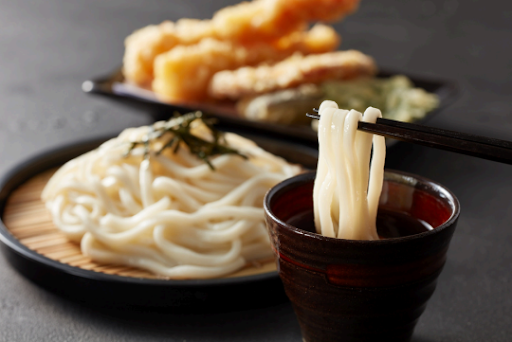 Chopsticks holding udon noodles over dipping sauce with tempura in the background on dark table. Noodles garnished with seaweed. Zaru udon noodles.