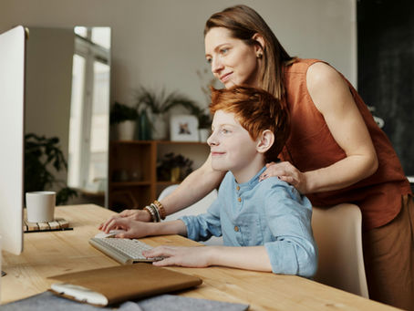 Mother gently supporting her red-haired son as he uses a desktop computer, both smiling in a warmly lit home office setting.