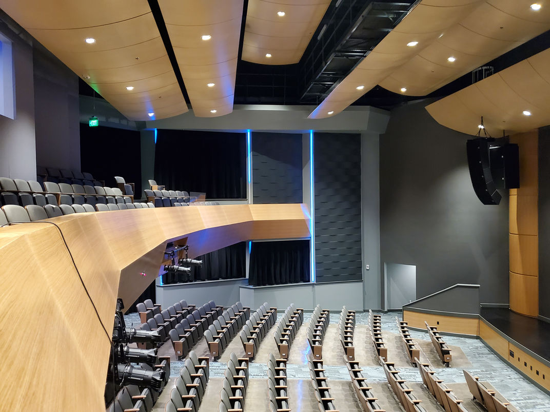 Interior view of a newly constructed auditorium with curved balcony seating, integrated wood finishes, acoustic wall treatments, and ceiling systems; building includes sound isolation and vibration control for HVAC.