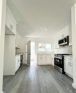 Long galley-style white kitchen with shaker cabinets, stainless steel appliances, and gray wood flooring.