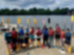 Group of paddlers in life jackets and holding paddles at a boat launch near the Connecticut River with trees in the background.