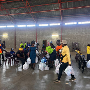 People in a large hall distribute and receive bags. Some wear bright shirts and hats. The mood is busy, with brick walls and a high ceiling.