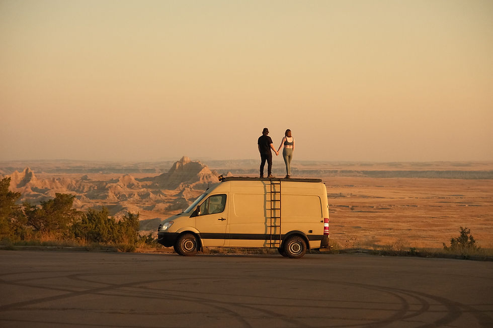 Two people stand holding hands on a van roof, overlooking a vast desert landscape at sunset. The scene feels serene and adventurous.