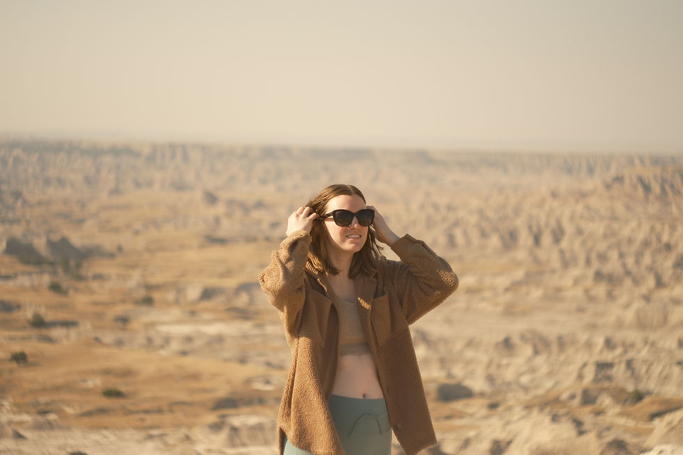 Woman in sunglasses smiling, wearing a brown jacket, standing in a vast desert landscape under a clear sky.