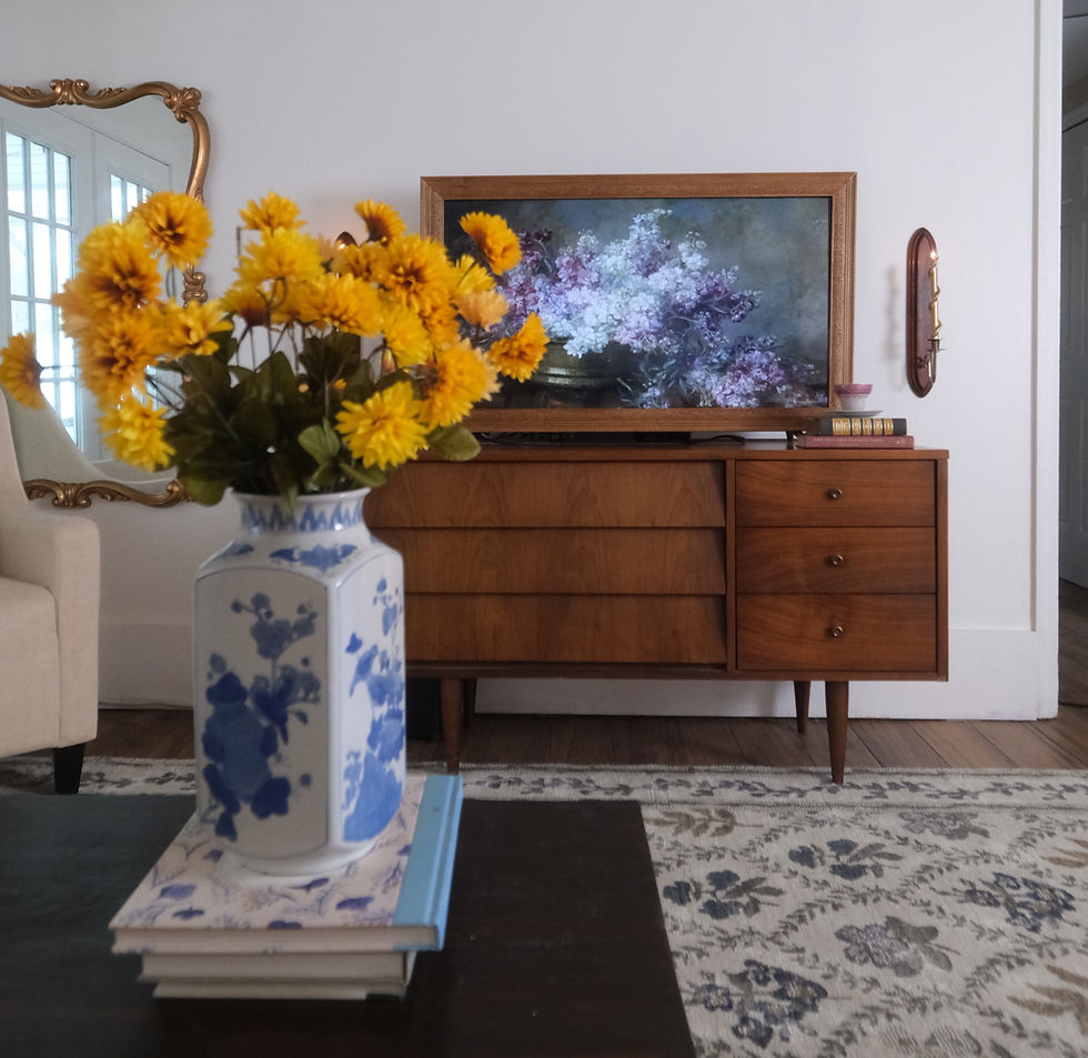 Vase with yellow flowers on a table, floral TV art, wooden dresser, ornate mirror, and patterned rug in a cozy living room.