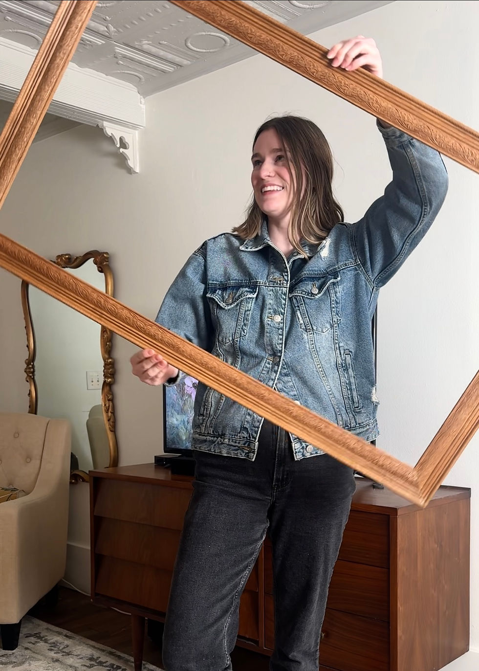 Smiling person in denim jacket holding a wooden frame in a vintage room with a mirror, chair, and wooden cabinet. Bright and cheerful mood.
