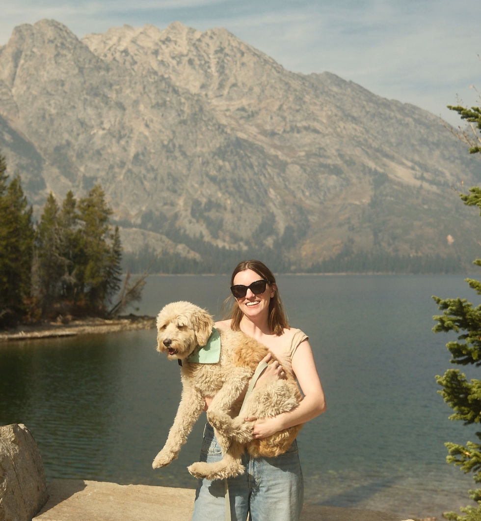 A woman in sunglasses holds a fluffy dog by a lake, with mountains and trees in the background. Both look content and relaxed.