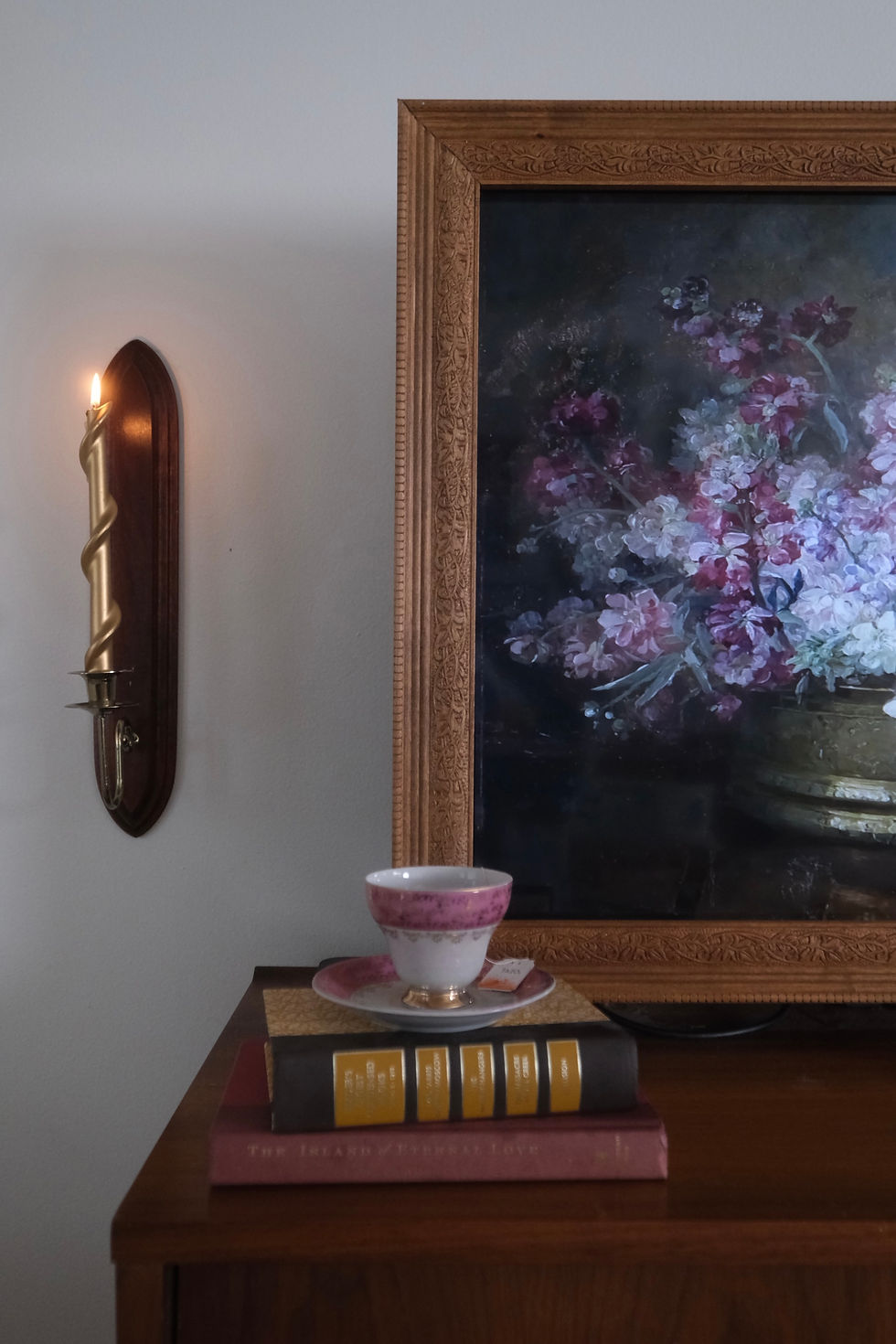Ornate painting of flowers next to a lit candle sconce. Pink teacup and saucer atop stacked books on a wooden table. Cozy ambiance.