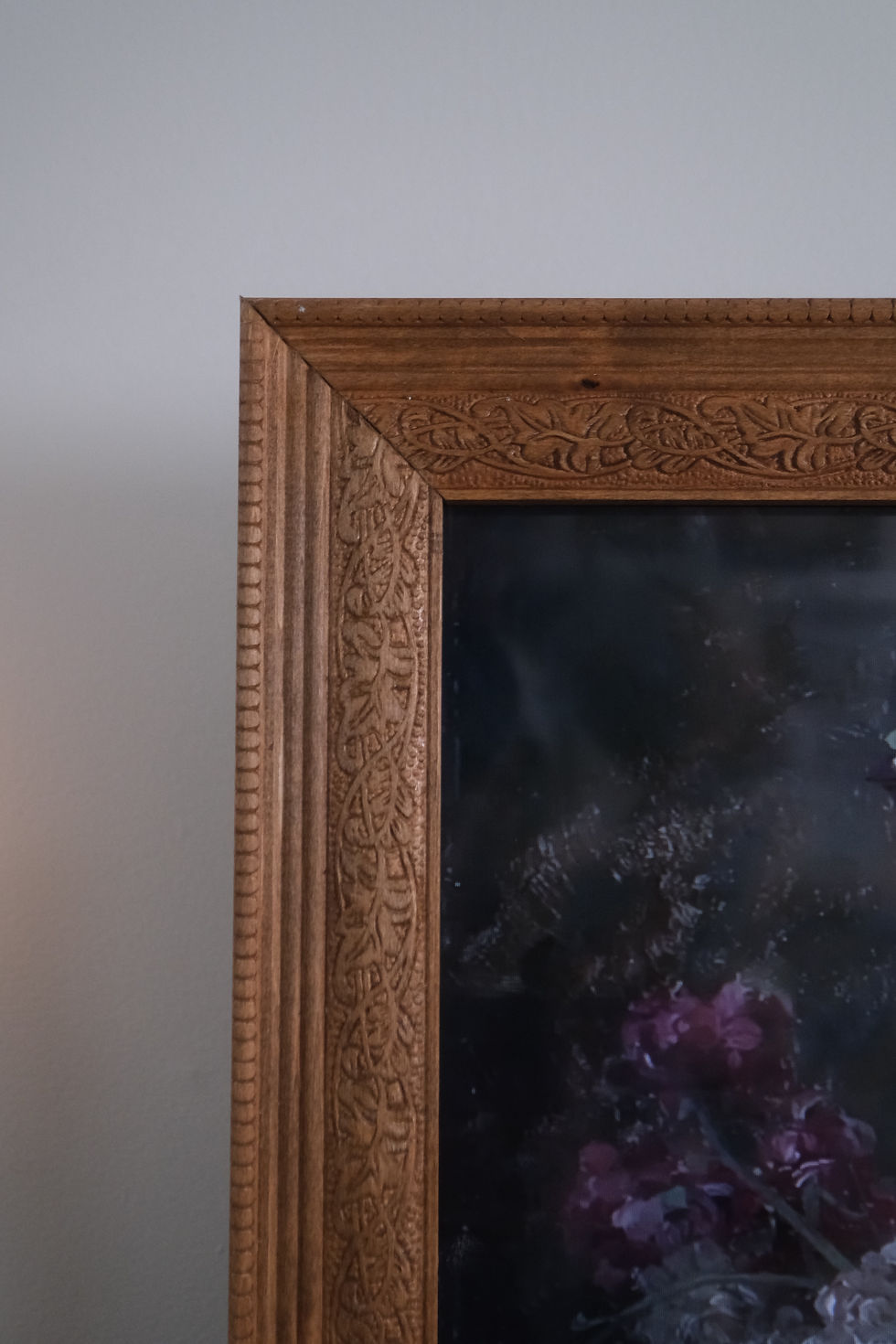 Close-up of a detailed wooden picture frame corner with floral carvings, set against a plain gray wall. Dark, moody artwork visible inside.
