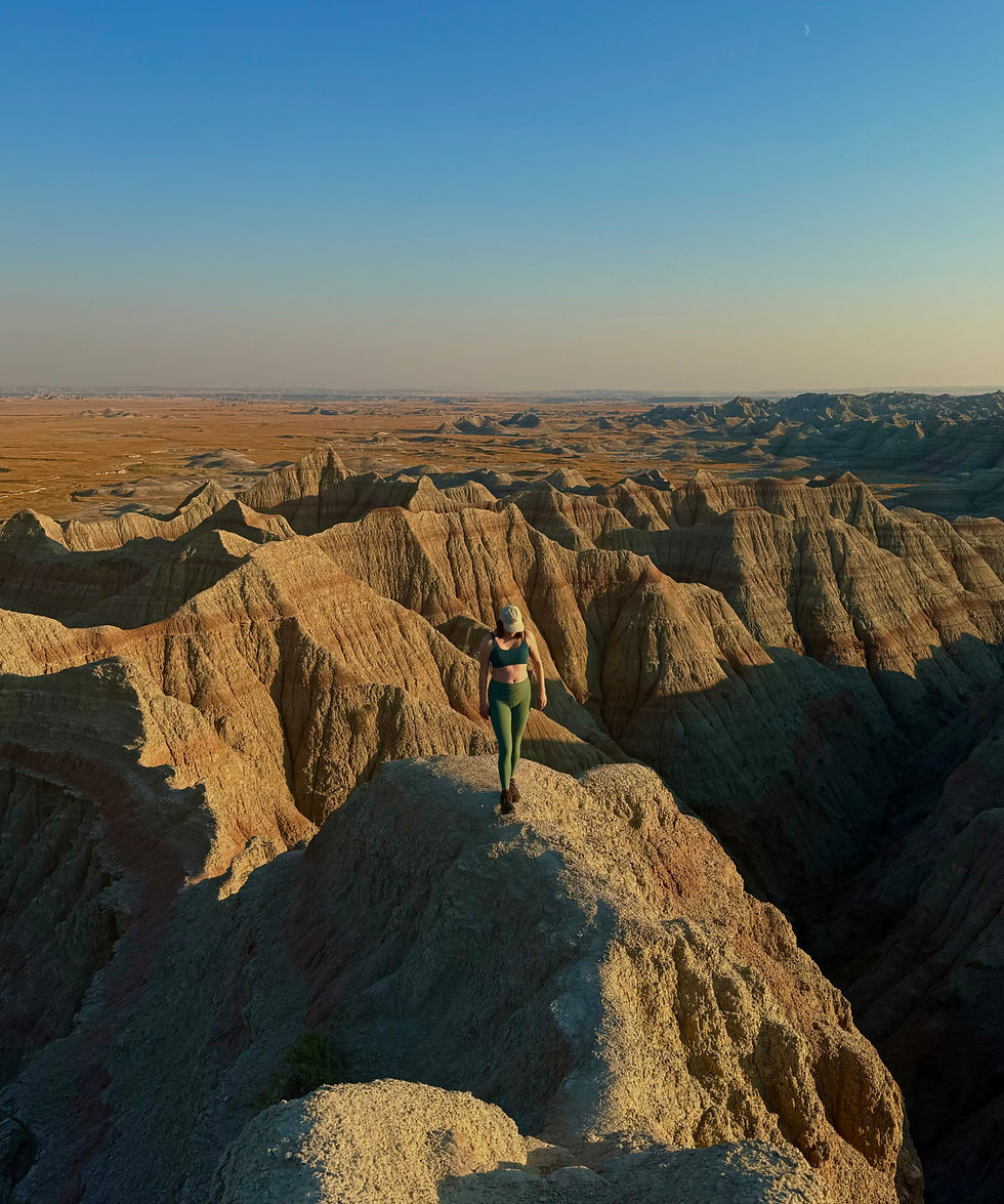 Person in athletic gear walks on rugged cliff edge at sunset in a vast, rocky desert landscape. Clear sky and warm tones set a serene mood.