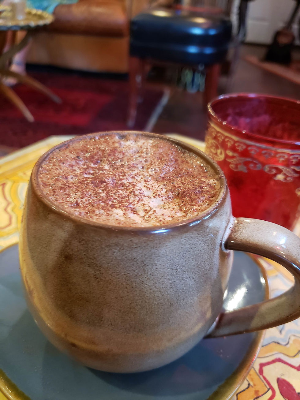 A close-up of a ceramic mug filled with frothy hot cacao, lightly dusted with cinnamon, sitting on a saucer atop a patterned table with a red glass and blurred background.