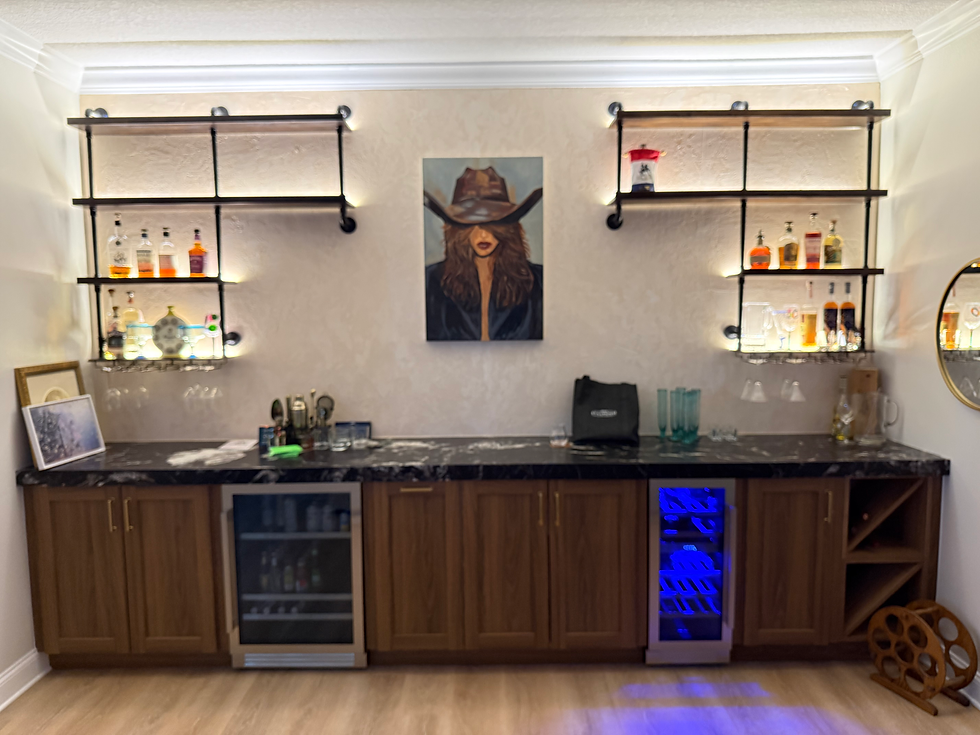 Stylish home bar with wood cabinets, liquor shelves, a cowboy portrait, glassware, plants, and warm lighting in a cozy living room