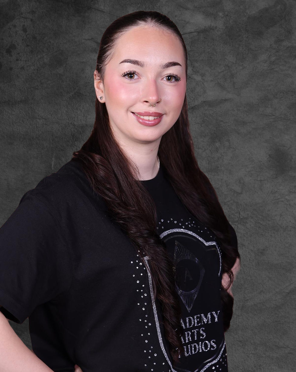 Kelsey, menyw ifanc yn gwisgo siwt du yn gwenu at y camera; Stiwdios Academy Arts.