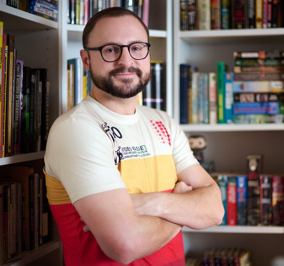 Writer Director Christian Rousseau in a square portrait, wearing a white and yellow video tape T-shirt standing in from of a book sheld, photographerd by Hart Getzen Photography.