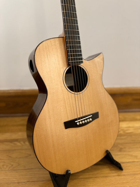 A guitar with a Florentine cutaway, sitka spruce top, ebony bridge and unbleached bone saddle and bridge pins sitting on a guitar stand.  The base side of the body is East Indian Rosewood and there is a soundport in the upper bout, base side. 