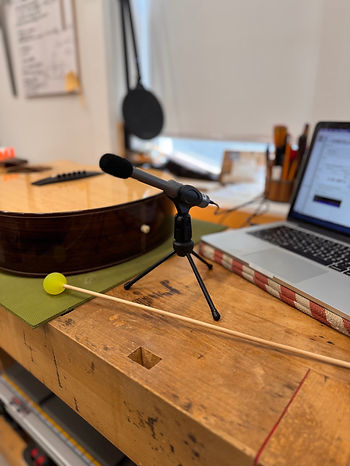 A microphone is mounted in a small stand and set ontop of a workbench. To its left is a finished guitar, to its right an open laptop and placed in front of the microphone is a small rubber mallet.