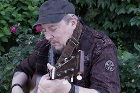 A close up on Singer/Songwriter Trace Ellis. He's wearing a black, worn baseball cap, a dark, rugged jeans jacket, a black and white leapord-print, thin scarf, and a gray t-shirt. He has a medallion around his neck. He's playing a guitar.