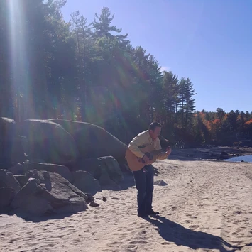 Singer/Songwriter Trace Ellis performing on a beach. He's wearing a light-colored long-sleeve shirt and playing. guitar. 