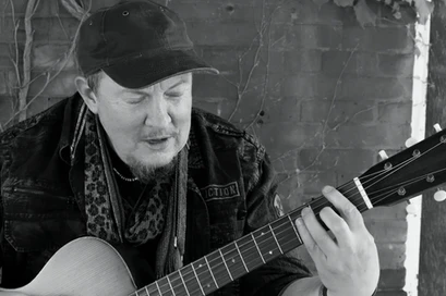A close up on Singer/Songwriter Trace Ellis. He's wearing a black, worn baseball cap, a dark, rugged jeans jacket, a black and white leapord-print, thin scarf, and a gray t-shirt. He has a medallion around his neck. He's playing a guitar. 