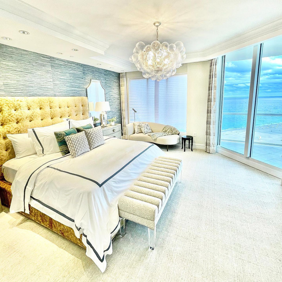 Luxury custom primary bedroom with floor-to-ceiling windows overlooking the ocean, featuring a modern upholstered bed, soft neutral tones, elegant lighting, porcelain tile flooring, and expansive coastal water views in a high-end Palm Beach County waterfront home.