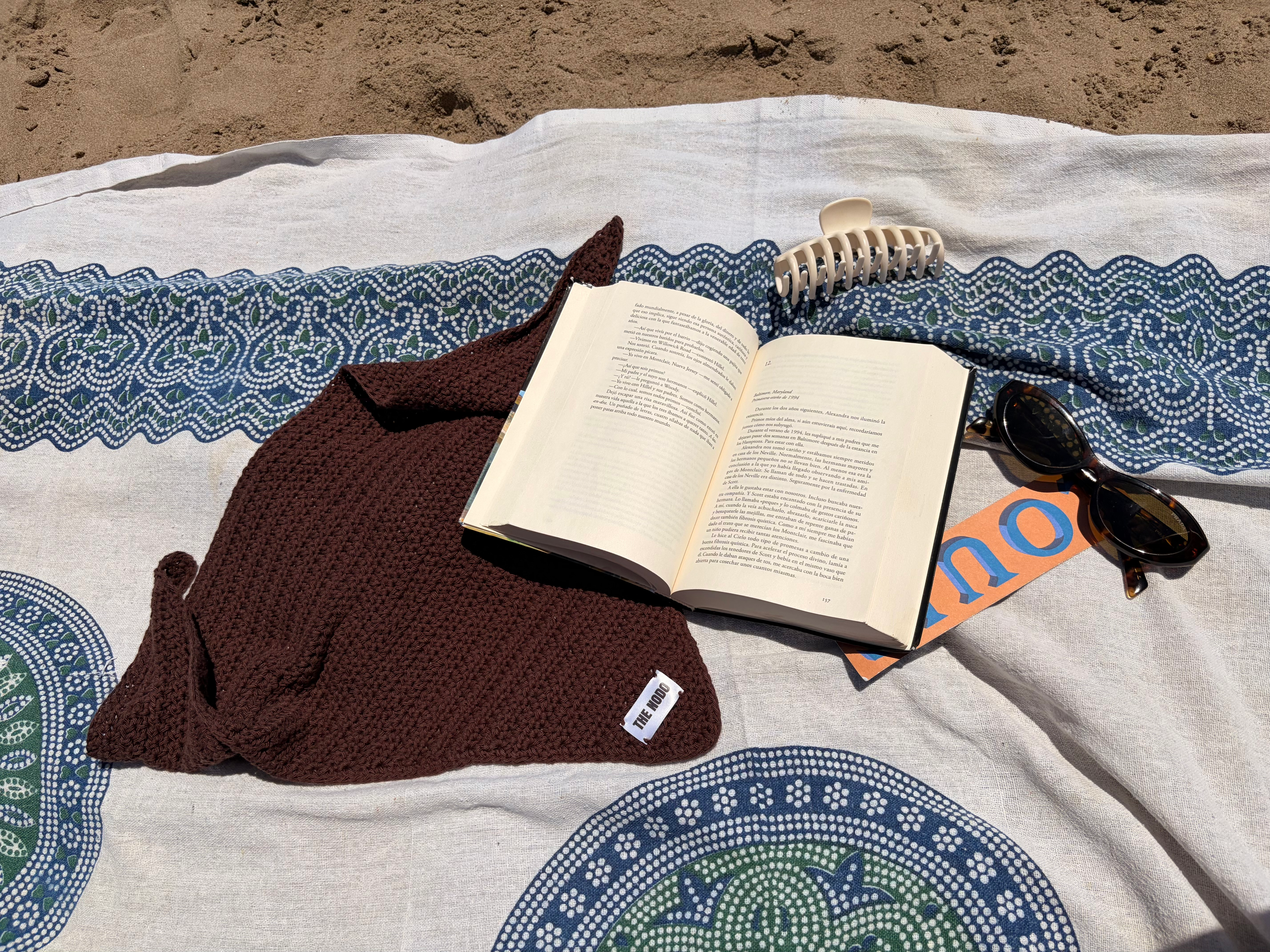 Bandana de punto marrón junto a un libro abierto sobre la arena de una playa española
