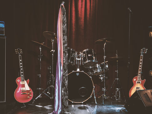 A stage with 2 electric guitars and a drum set backed by a burgundy curtain