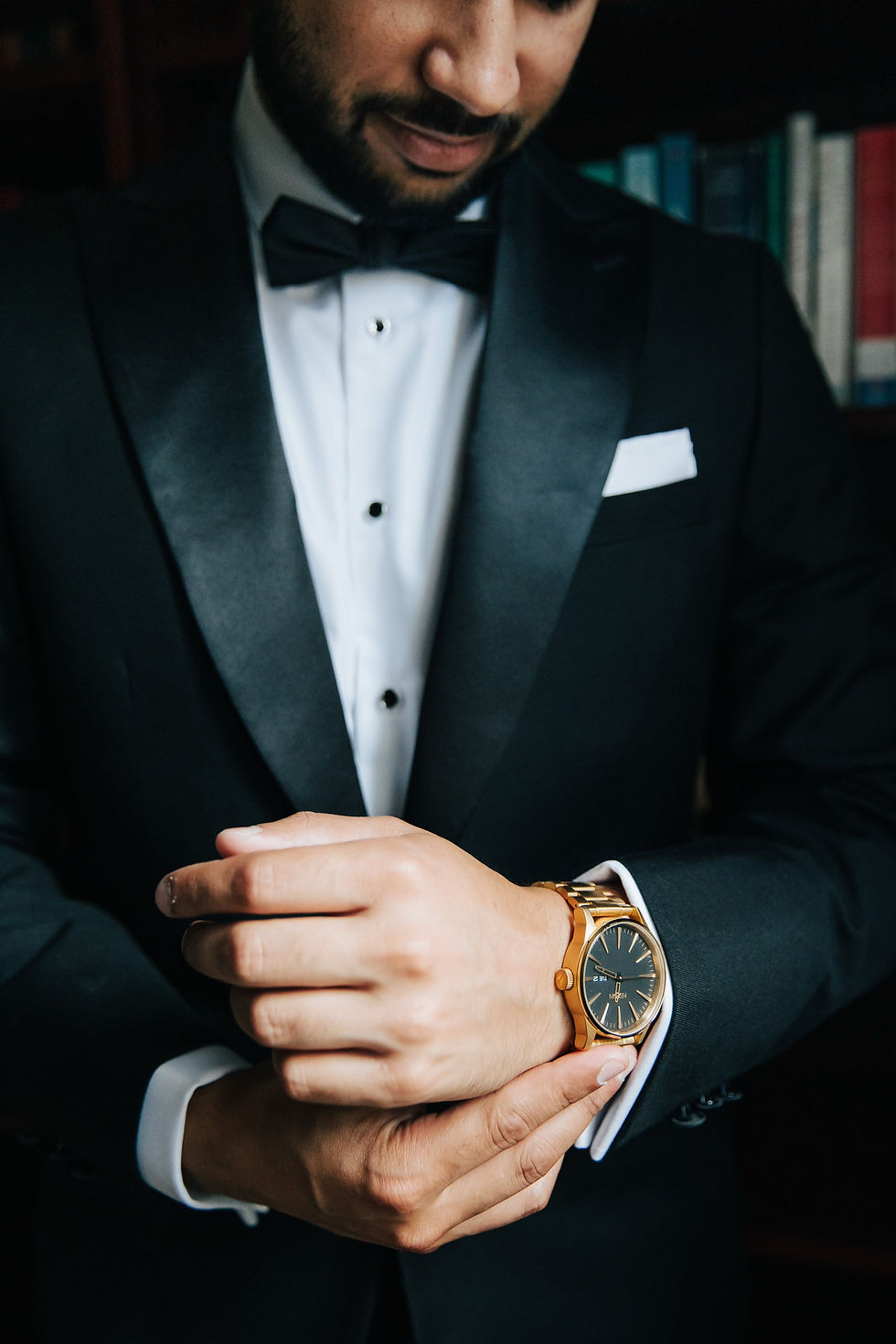 Man in a black tuxedo adjusts his gold watch. Background features blurred bookshelves. Mood is formal and composed.