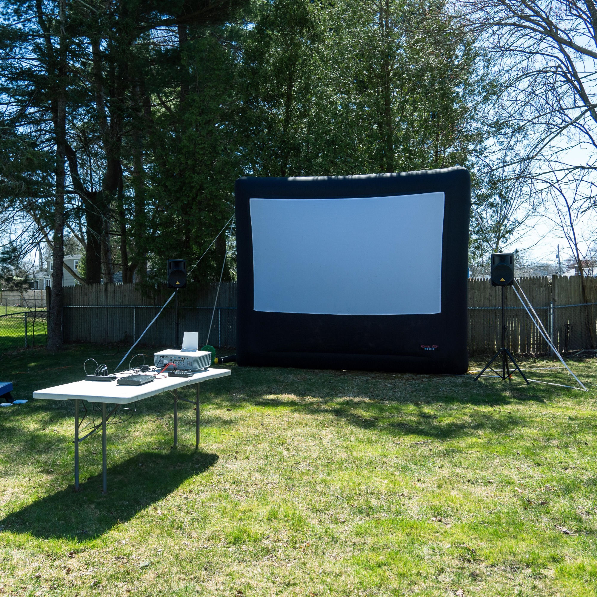 a 16 foot inflatable screen with a table pointing at it with a projector on top of it there is a cornhole board on the left. 