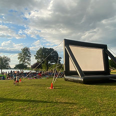 a GIANT inflatable movie screen about 3 stories high. Children and adults scattered in front of it waiting for the sun to set and the movie to begin (reverse angle)