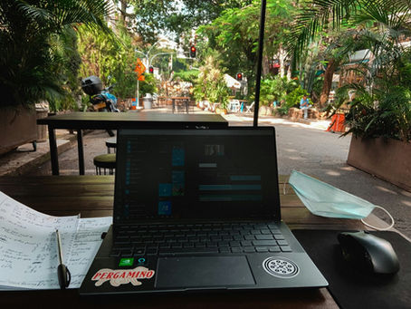 Photo d’un poste de travail en plein air avec un ordinateur portable, des notes manuscrites et un masque chirurgical sur une table en bois. La scène se déroule dans une rue végétalisée et calme, avec des tables de café en arrière-plan, idéale pour le télétravail. L’environnement verdoyant, les palmiers et la lumière naturelle créent une ambiance relaxante et productive.