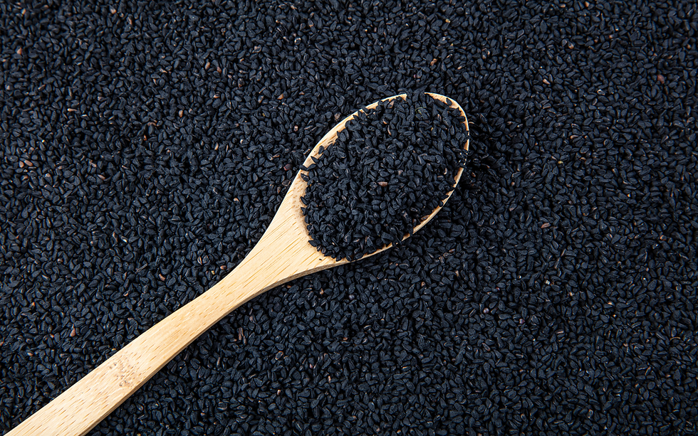 Das Bild zeigt einen hölzernen Löffel, der mit Schwarzkümmelsamen gefüllt ist, die sich nahtlos in einen Hintergrund aus vielen weiteren Schwarzkümmelsamen einfügen. Die kleinen, schwarzen Samen sind dicht angeordnet, und der Löffel hebt sich mit seiner natürlichen Holzfarbe kontrastreich von den dunklen Samen ab. Die Textur der Samen ist rau und körnig, was die natürliche Beschaffenheit des Schwarzkümmels betont.