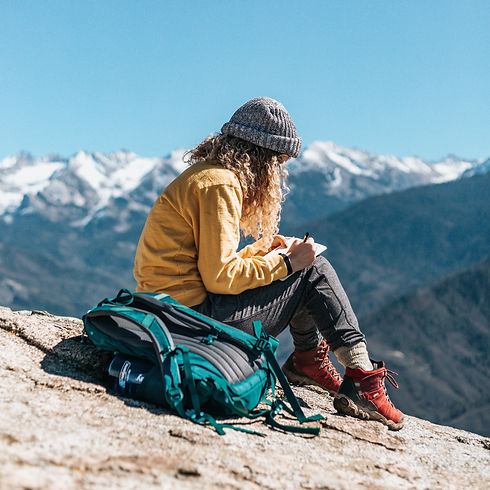Woman writing in notebook, sitting on rock, mountain backdrop and blue sky.