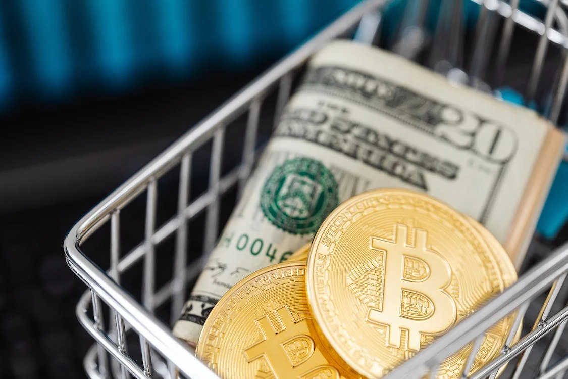 Beginner's Guide to Buy Bitcoins: How to Get Started with Bitcoin Trading" article featured image of a person using a laptop to purchase bitcoins with a mobile phone and a physical bitcoin on the desk.