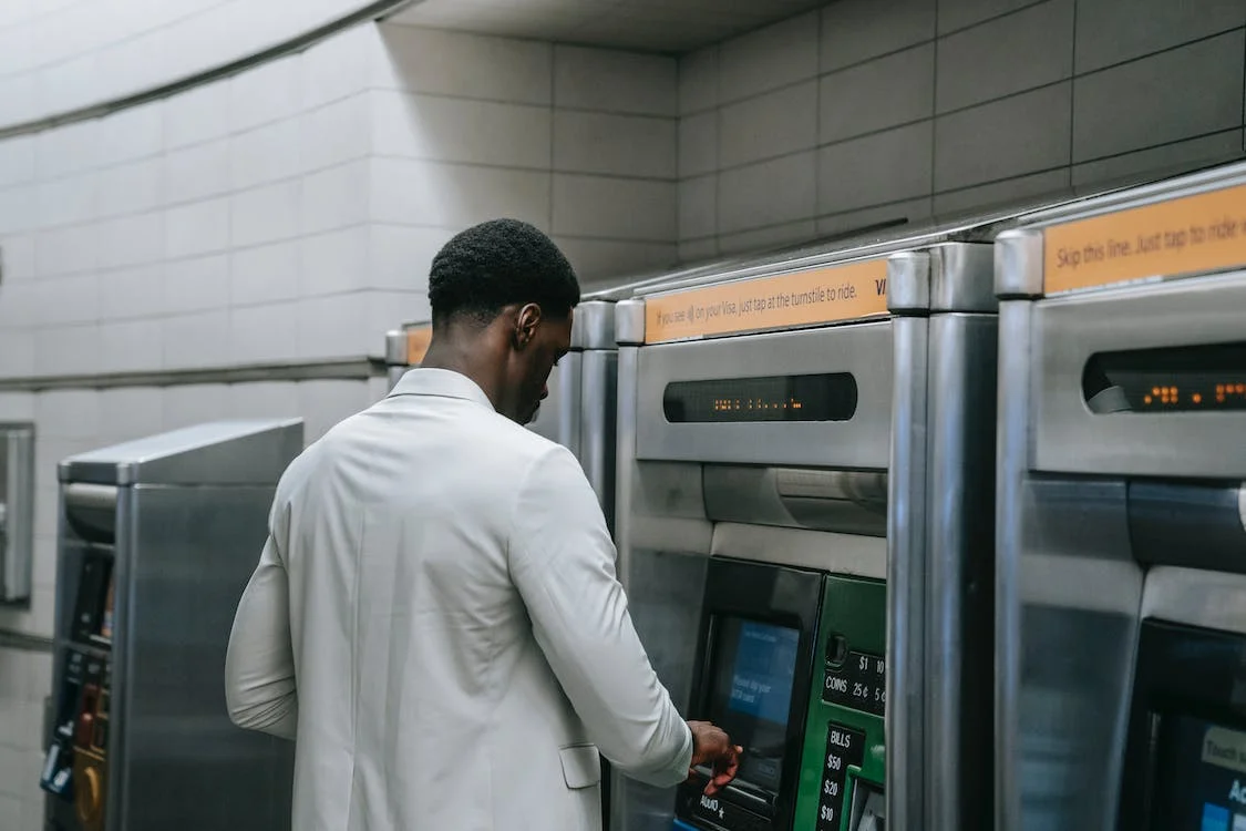An ATM machine with a screen displaying a Bitcoin logo and a QR code. The machine is located in a well-lit indoor area with a seating arrangement nearby. People can be seen walking in and out of the area in the background. The image represents a Bitcoin ATM in Georgia that allows users to buy Bitcoins easily.