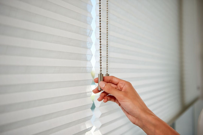 Hånd trekker i snor for å justere hvite plissegardiner i et hus.
