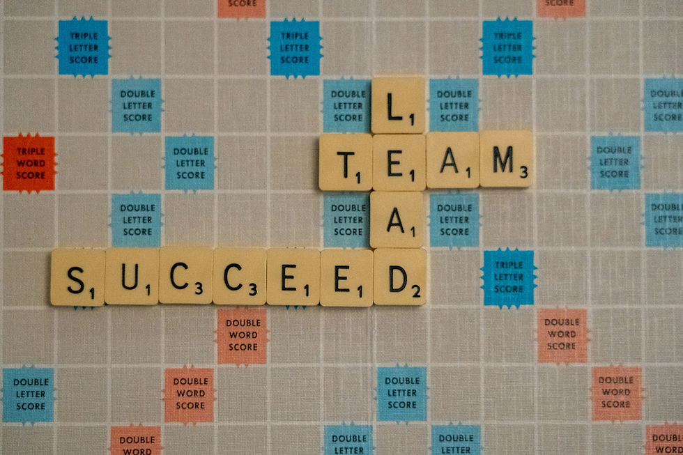 Scrabble board with tiles forming "TEAM," "LEAD," and "SUCCEED." Background has blue and red score labels, conveying teamwork and success.