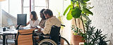A team working in a brightly lit office with natural lighting, large plants and design accommodations for an employee in a wheelchair.