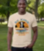 Natural unisex t-shirt worn by smiling man in park, "Be Kind To Every Kind" cow fish sunset retro graphic.