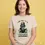 Thumbnail: Natural t‑shirt with cartoon Darth Vader parody holding cauliflower and broccoli, worn by woman in casual pose.