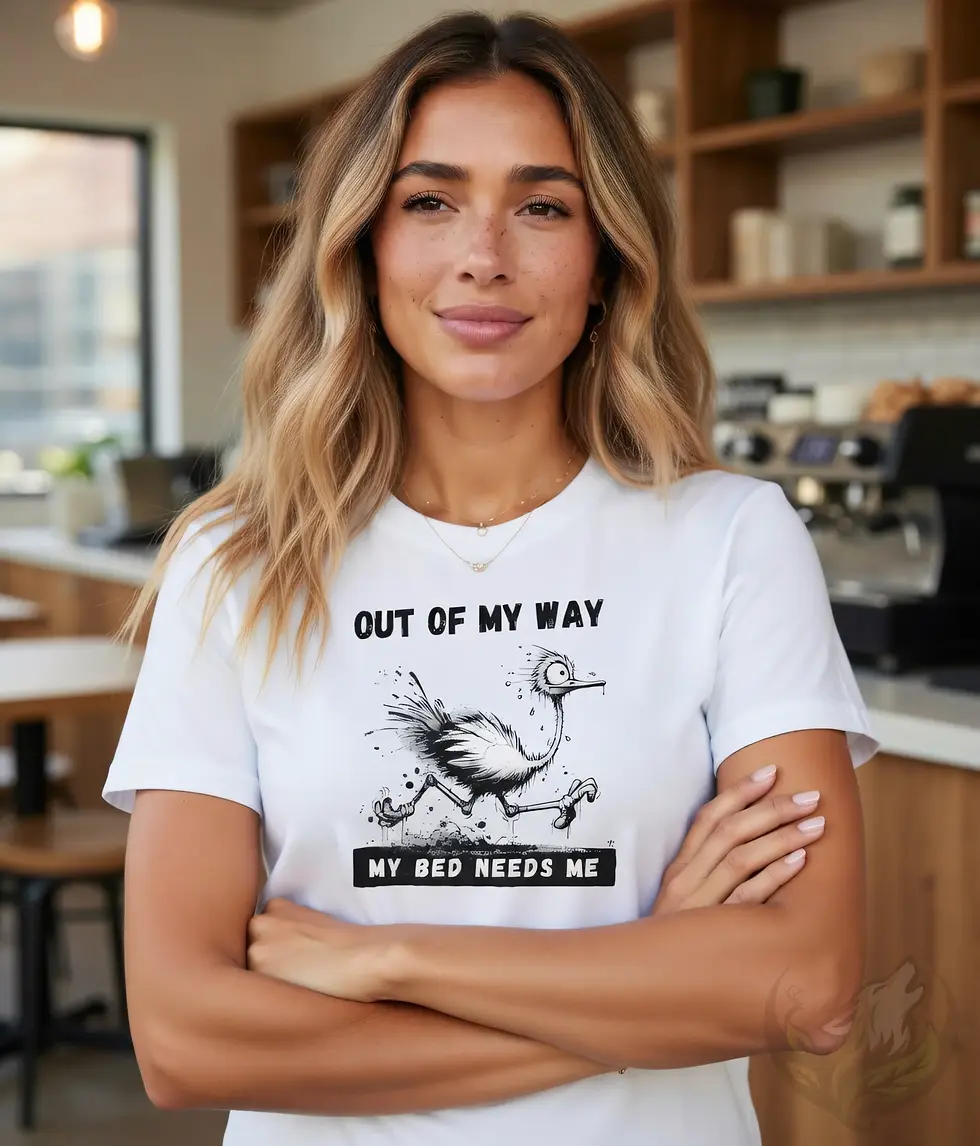Thumbnail: Smiling woman standing in a kitchen wearing a white fitted t-shirt featuring the humorous "My Bed Needs Me" running ostrich d