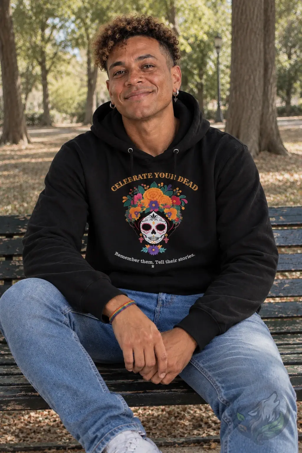 Man wearing a black Celebrate Your Dead hoodie with colourful sugar skull design on a park bench