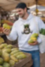 Man wearing a white unisex hoodie with an angry graffiti pineapple design at a fruit market.