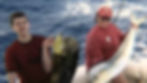 A photograph of a young man and a woman on a boat in the ocean, both smiling and holding large fish they have caught. The man is on the left holding a dark-colored fish, and the woman is on the right holding a large, lighter-colored mahi-mahi.