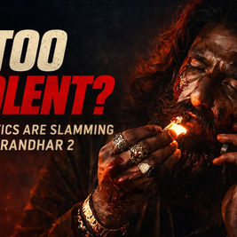A cinematic 16:9 promotional image featuring a bloodied man with long hair and a beard lighting a cigarette with a match. The scene uses dark tones with red highlights and floating embers to create an intense atmosphere. Bold text on the left reads “TOO VIOLENT?” with a subheading referencing Dhurandhar 2 and criticism from reviewers.
