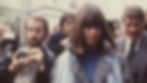 A medium shot of Marianne Bachmeier in the early 1980s, walking through a crowd of people. She has shoulder-length brown hair with bangs and is wearing a grey trench coat over a blue top with a light blue scarf. She is looking down with a somber expression while being surrounded by photographers and men in suits, suggesting a high-profile legal or media event.