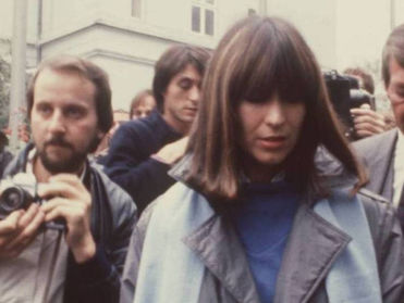 A medium shot of Marianne Bachmeier in the early 1980s, walking through a crowd of people. She has shoulder-length brown hair with bangs and is wearing a grey trench coat over a blue top with a light blue scarf. She is looking down with a somber expression while being surrounded by photographers and men in suits, suggesting a high-profile legal or media event.