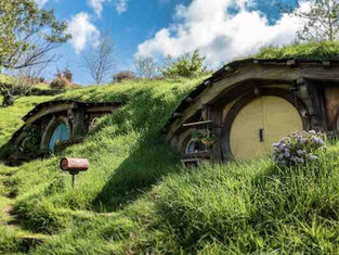 Two Hobbit Holes built into a grassy hillside, with their iconic round doors visible. The one on the right has a light yellow door, and the one on the left has a blue door. A small, rustic mailbox on a post sits between them. A narrow dirt path with stone steps leads up the hill under a bright, sunny sky