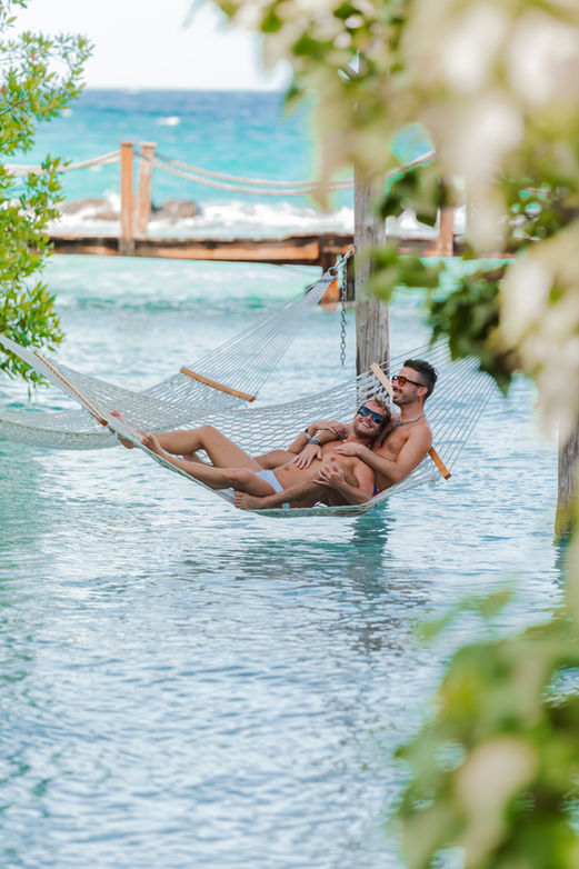 Gay couple relaxing together on a hammock at Renaissance Island Aruba over the water at a tropical beach, captured by Edrxck, an Aruba photographer specializing in LGBTQ+ vacation photography.